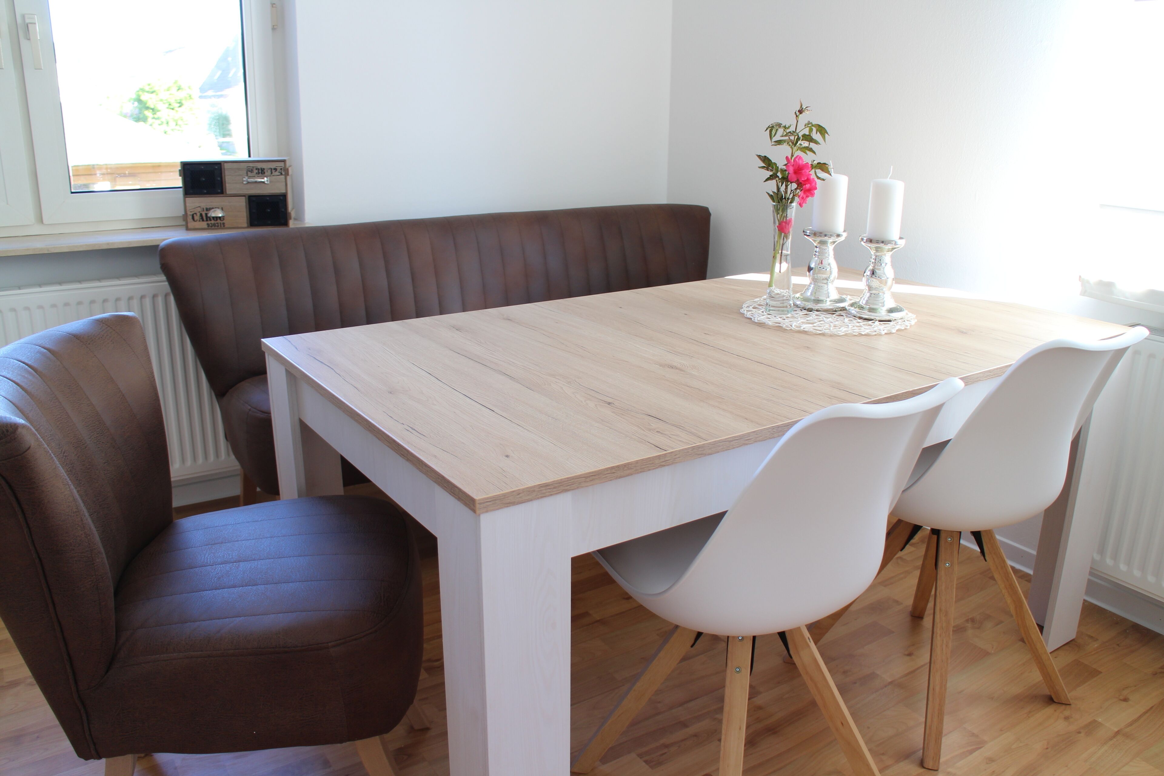 Un coin repas avec une table en bois, des chaises blanches, un banc marron et un vase orné de fleurs et de bougies.
