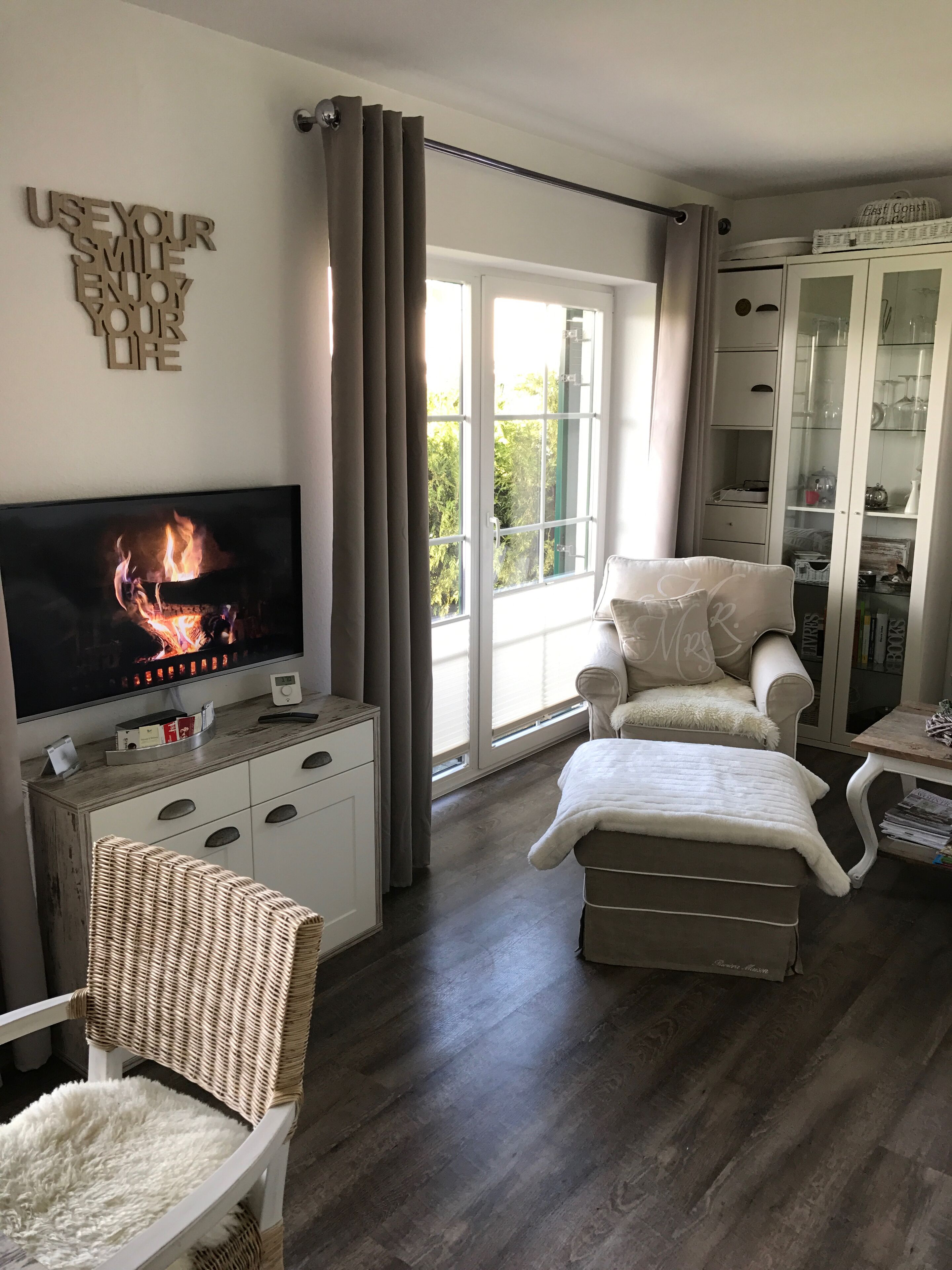 Ein gemütliches Wohnzimmer mit Fernseher, der einen Kamin zeigt, einer bequemen Couch, einem Korbsessel und einer Vitrine mit Regalen.