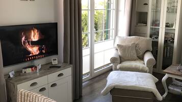 Ein gemütliches Wohnzimmer mit Fernseher, der einen Kamin zeigt, einer bequemen Couch, einem Korbsessel und einer Vitrine mit Regalen.