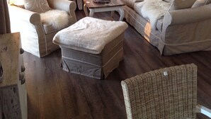 A cozy living room with a sofa set, a chair, a small ottoman, and a wicker chair. There is a window with curtains, a radiator, and a hanging light fixture.