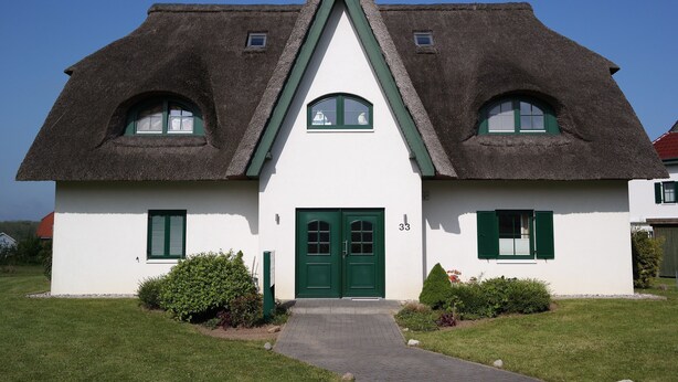 A traditional house with a steeply pitched thatched roof, a central entrance with a green door, and two windows with green shutters.
