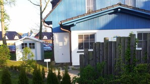 Una casa de dues plantes amb façana blava i teulat marró, envoltada d'una tanca de fusta i diferents plantes.