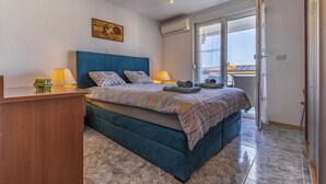 A bedroom with a bed, two bedside lamps, a wall-mounted air conditioner, and a window with a view of buildings.