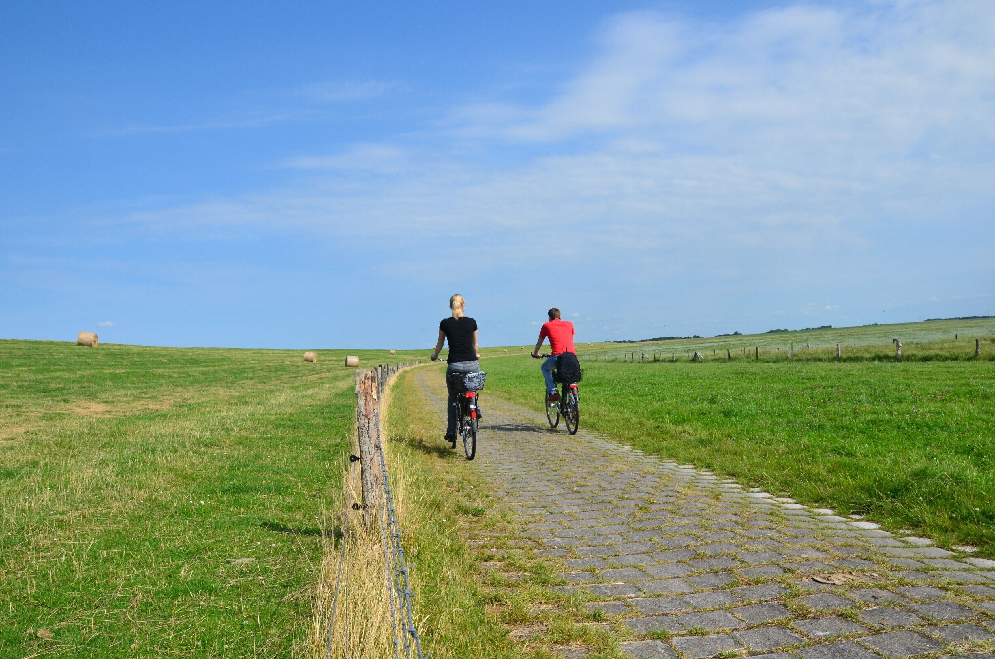 Deux personnes à vélo sur un chemin pavé de pierres, dans un champ verdoyant parsemé de bottes de foin, sous un ciel dégagé.
