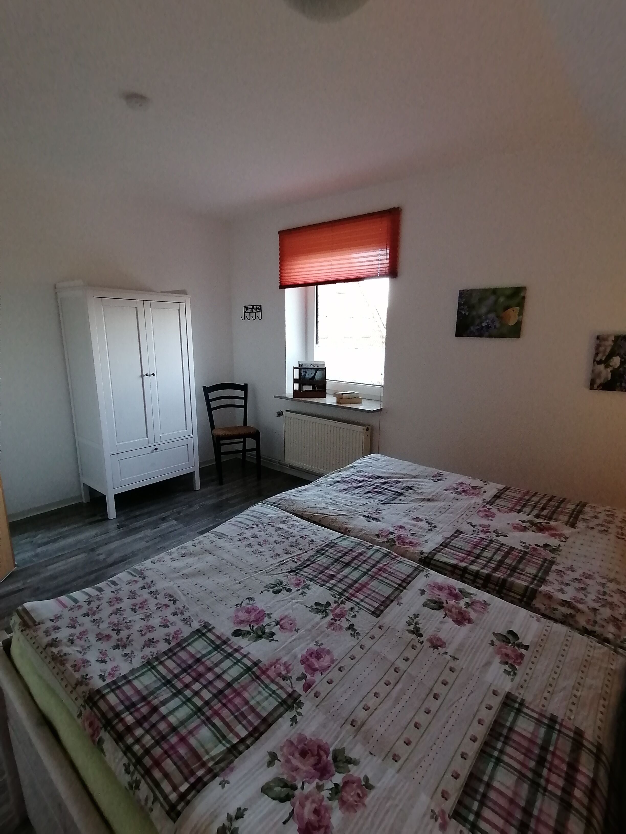 Una camera da letto con un letto, un armadio, una sedia, una finestra con un parasole rosso e un termosifone.