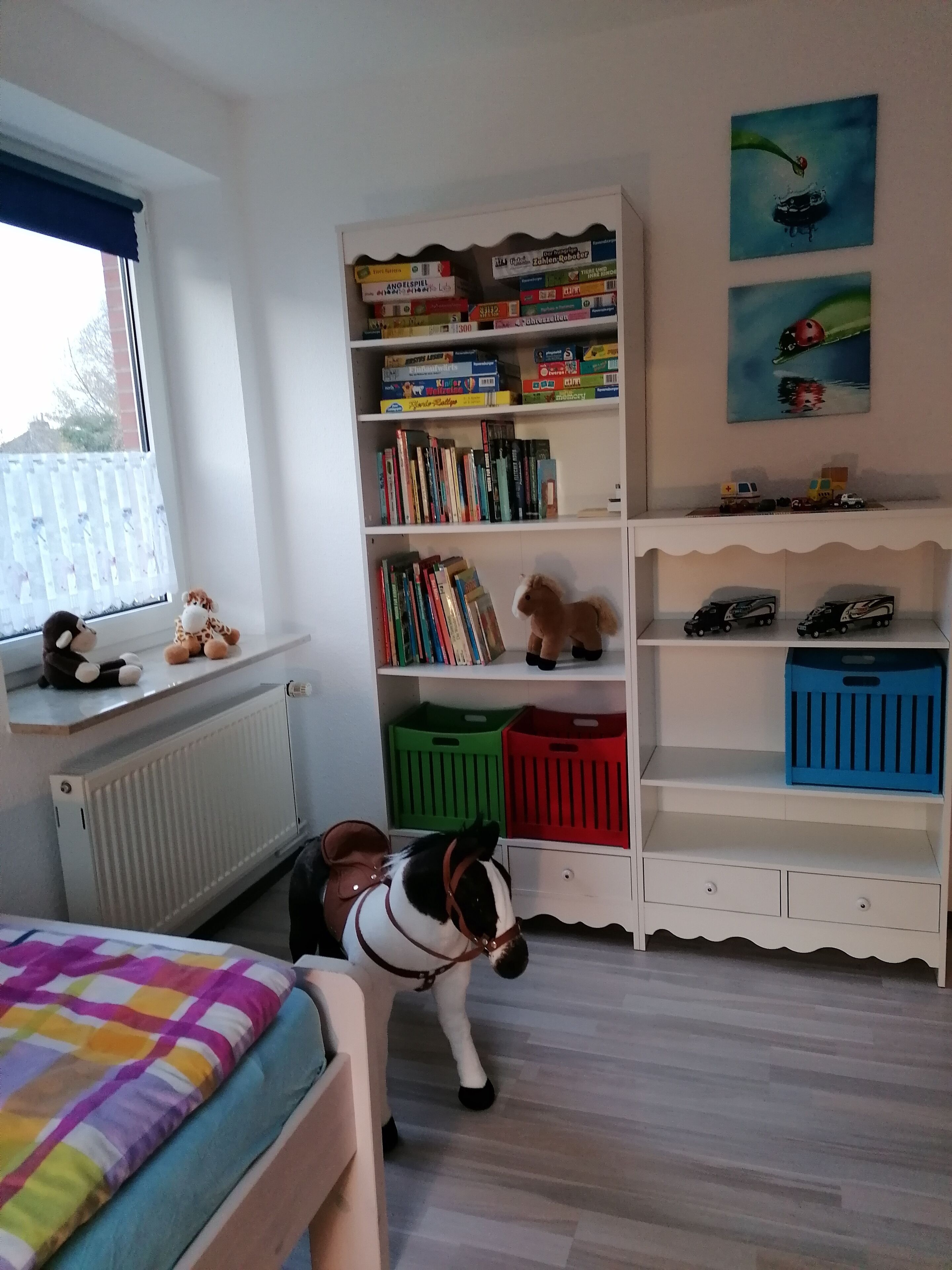 Ein Kinderzimmer mit Bücherregal, Heizkörper, Fenster mit Aussicht und einem Bett mit bunter Decke.
