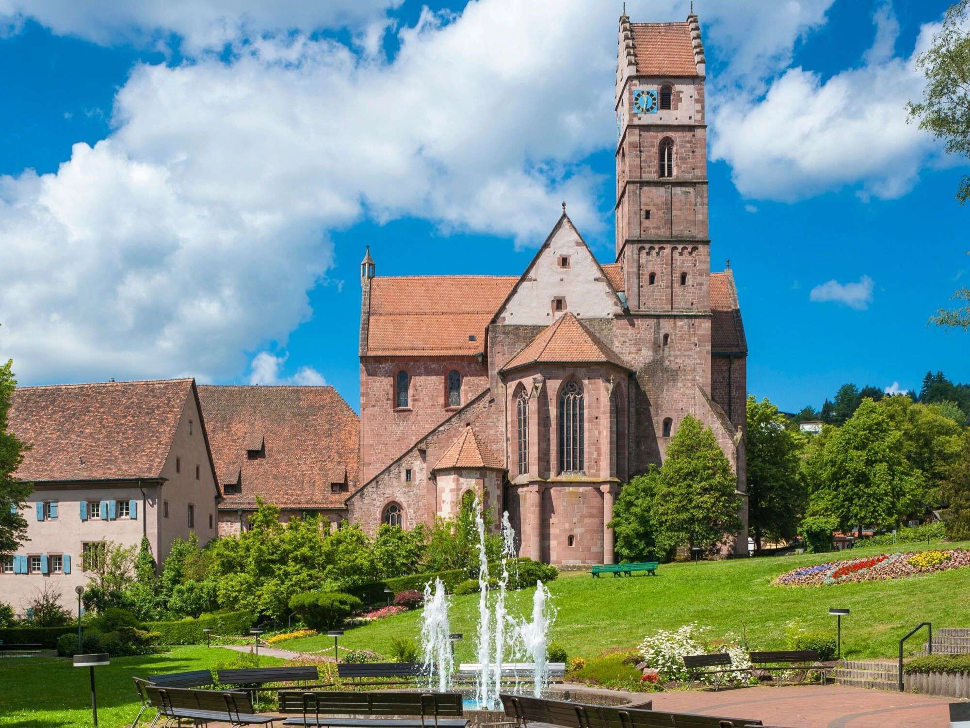 Eine historische Kirche mit einem markanten Glockenturm, umgeben von einem gepflegten Garten mit Brunnen, Bänken und einem Weg.