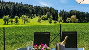 Ein Tisch mit zwei Stühlen, einer Champagnerflasche und einem Blumenstrauß, vor einer Kulisse aus einem grünen Feld und einem Wald.