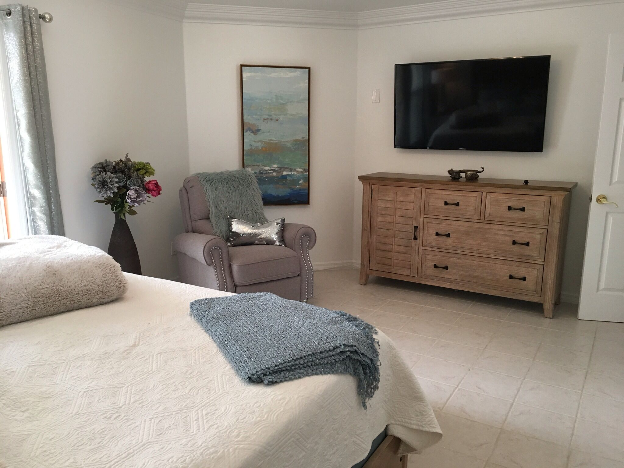 A bedroom with a bed, a chair, a dresser, a painting, and a TV.