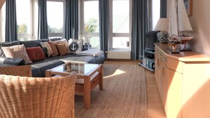 Een ruime woonkamer met een donkergrijze bank, een bijzettafeltje en een rieten fauteuil. Er staat een televisie op een dressoir, een klein tafeltje met een lamp en een boekenkast. Grote ramen met gordijnen zorgen voor veel daglicht.