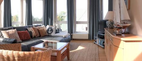 Ein geräumiges Wohnzimmer mit einem dunkelgrauen Sofa, einem Couchtisch und einem Korbstuhl. Ein Fernseher steht auf einem Schrank, ein kleiner Tisch mit Lampe und ein Bücherregal sind ebenfalls vorhanden. Große Fenster mit Vorhängen sorgen für natürliches Licht.