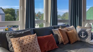 Een moderne woonkamer met een donker leren bank, meerdere kussens en een uitzicht op een groen landschap door grote ramen.