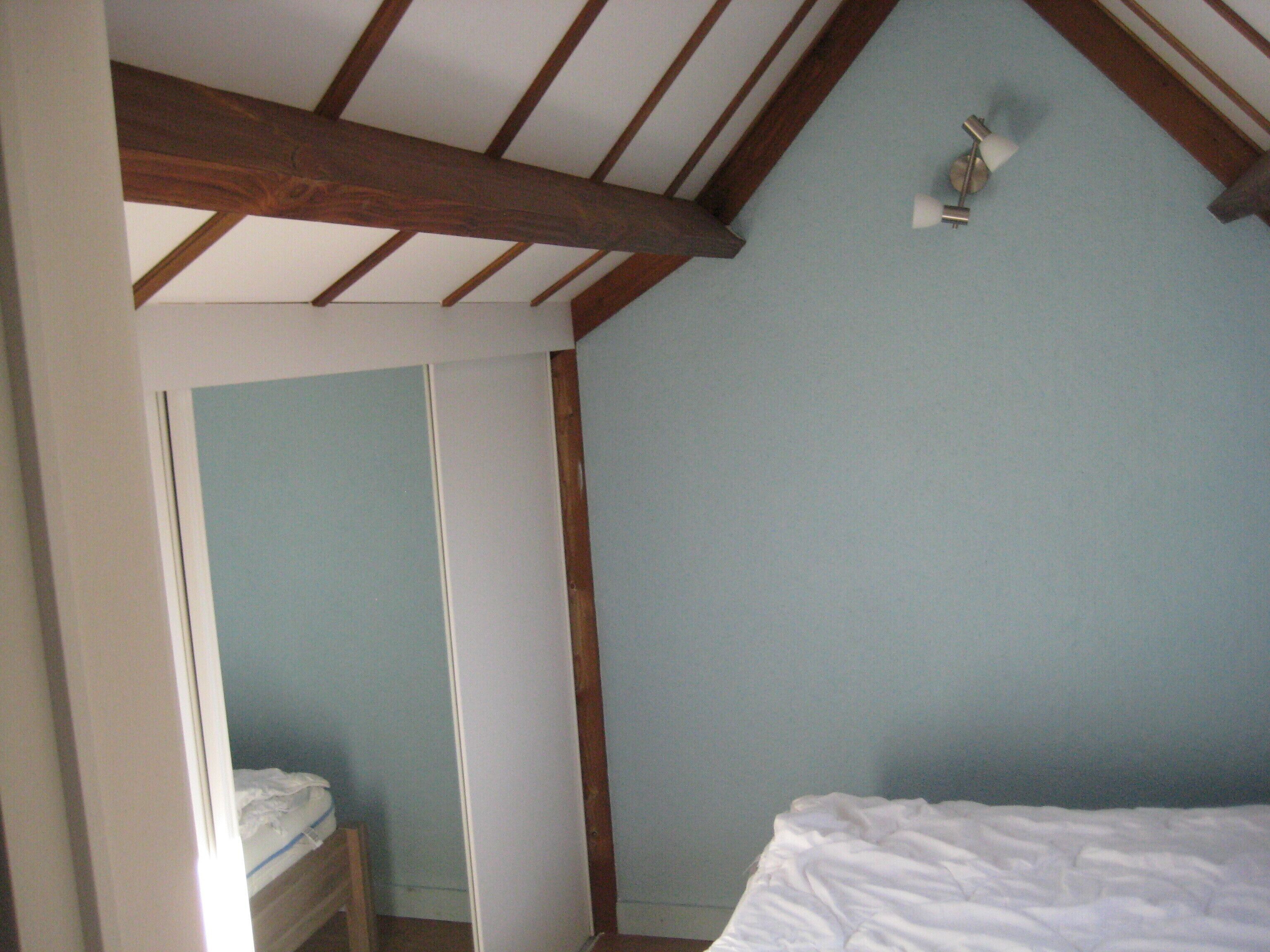 A bedroom with a sloped ceiling, wooden beams, a bed, a nightstand, and a wall-mounted light fixture.