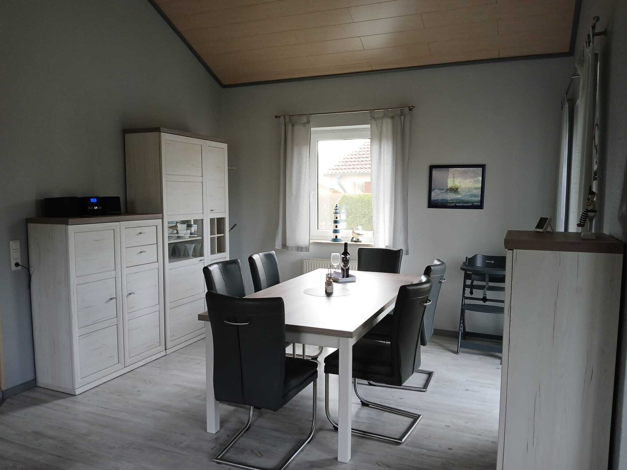 A compact kitchen with a dining table, chairs, a cabinet with drawers, a window with curtains, and a small shelf with a decorative item.