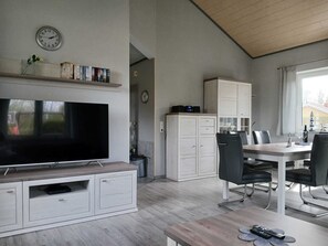 Uma sala de estar moderna com televisão de tela plana, um espaço de refeições com mesa e cadeiras, e uma cozinha com armários e um relógio.