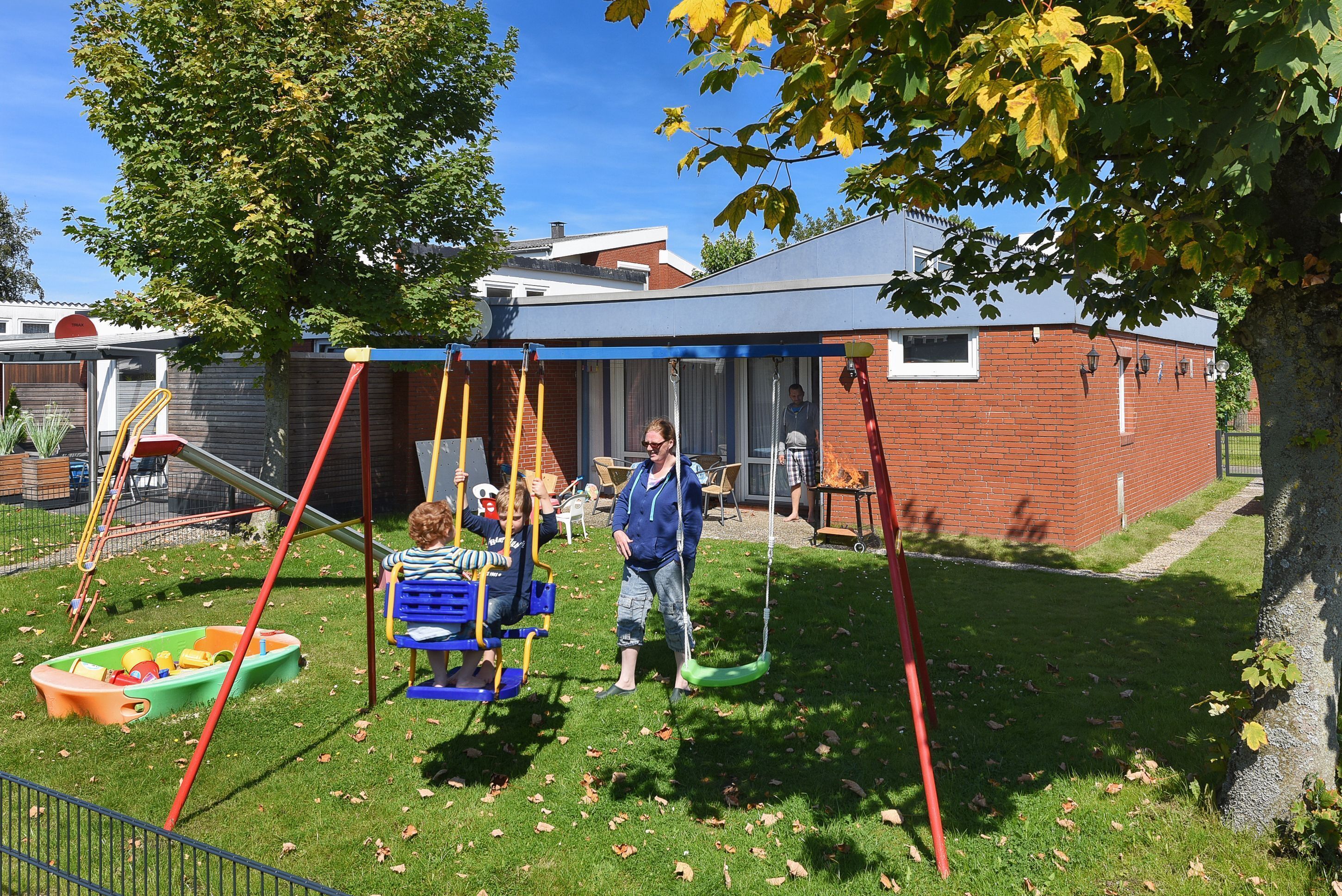 A playground with swings and slides, a brick building in the background, and a tree providing shade.