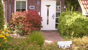 A brick building with a red-tiled roof, a white door, and a small garden with various plants and flowers. - Sunny spacious. DHH, 3 bedrooms, 6 pers., NR, WLan, close to the beach, washer + dryer (Niedersachsen)