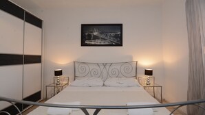 Une chambre à coucher bien rangée, avec un lit, deux tables de chevet dotées de lampes, un tableau encadré au mur et un rideau à la fenêtre.