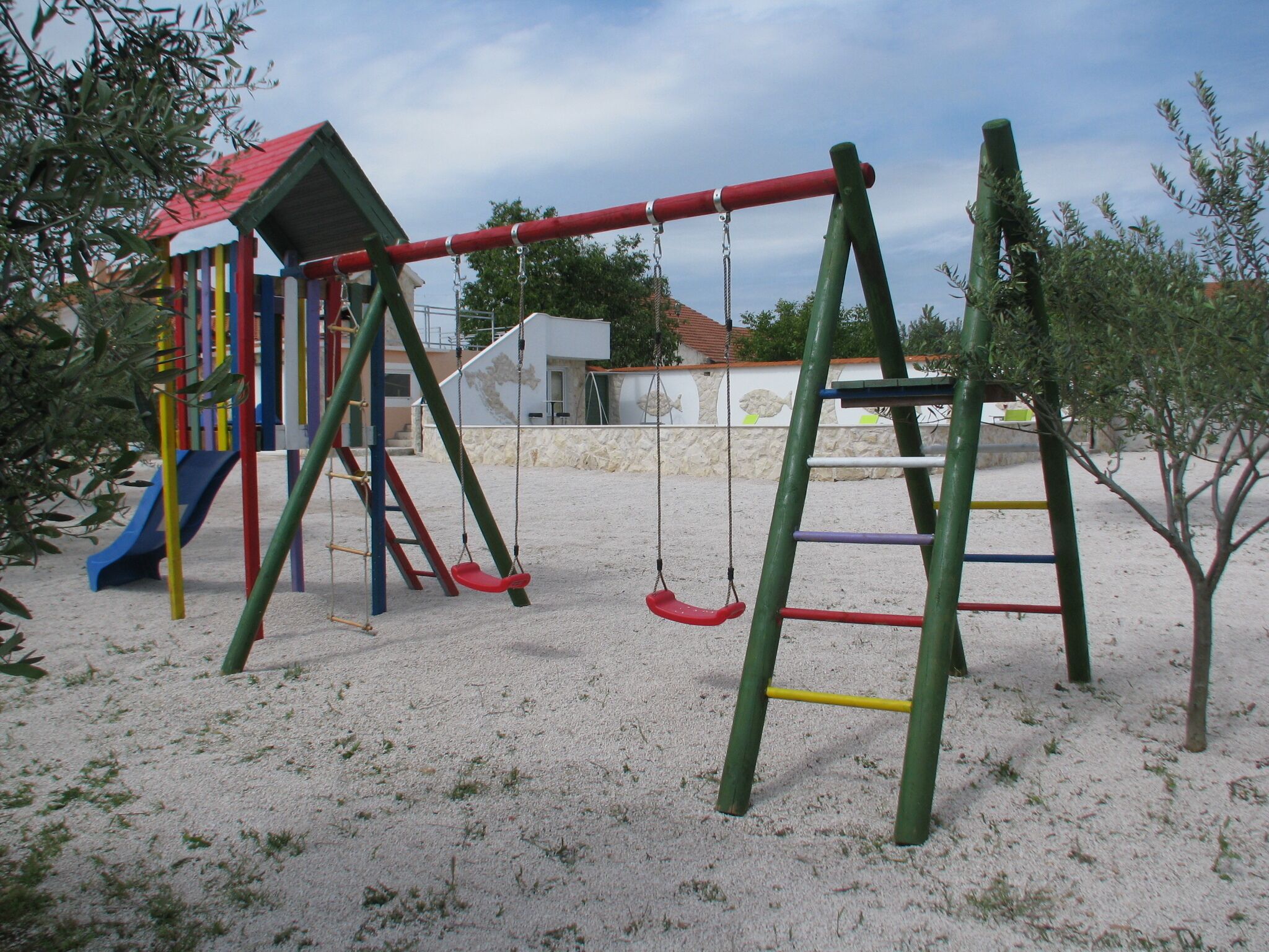 Un terrain de jeux avec des balançoires et un toboggan, entouré de sable et d’une végétation clairsemée.