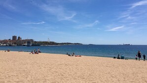 Una playa con gente tomando el sol y nadando, un cielo despejado y un paisaje urbano al fondo.