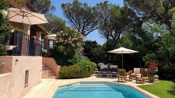 Zona de piscina con vista despejada, rodeada por un jardín con diversas plantas y árboles. Hay mobiliario exterior y sombrillas que brindan sombra.