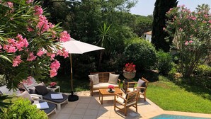 Zona junto a la piscina con una mesa y sillas, una sombrilla blanca, y vistas a un jardín exuberante y un edificio al fondo.