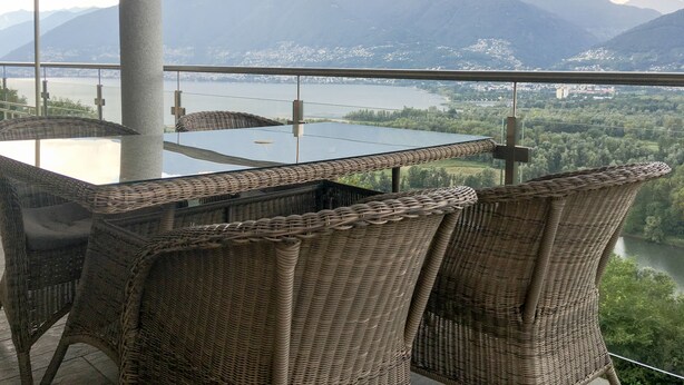 A balcony with wicker furniture overlooking a mountainous landscape and a body of water. - 4* Vacation Home Residenza Christa - Casa Wiro (Magadino - Gambarogno)