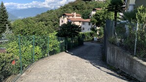 A narrow, paved road flanked by greenery and residential buildings with a mountainous backdrop. - 4* Vacation Home Residenza Christa - Casa Wiro (Magadino - Gambarogno)