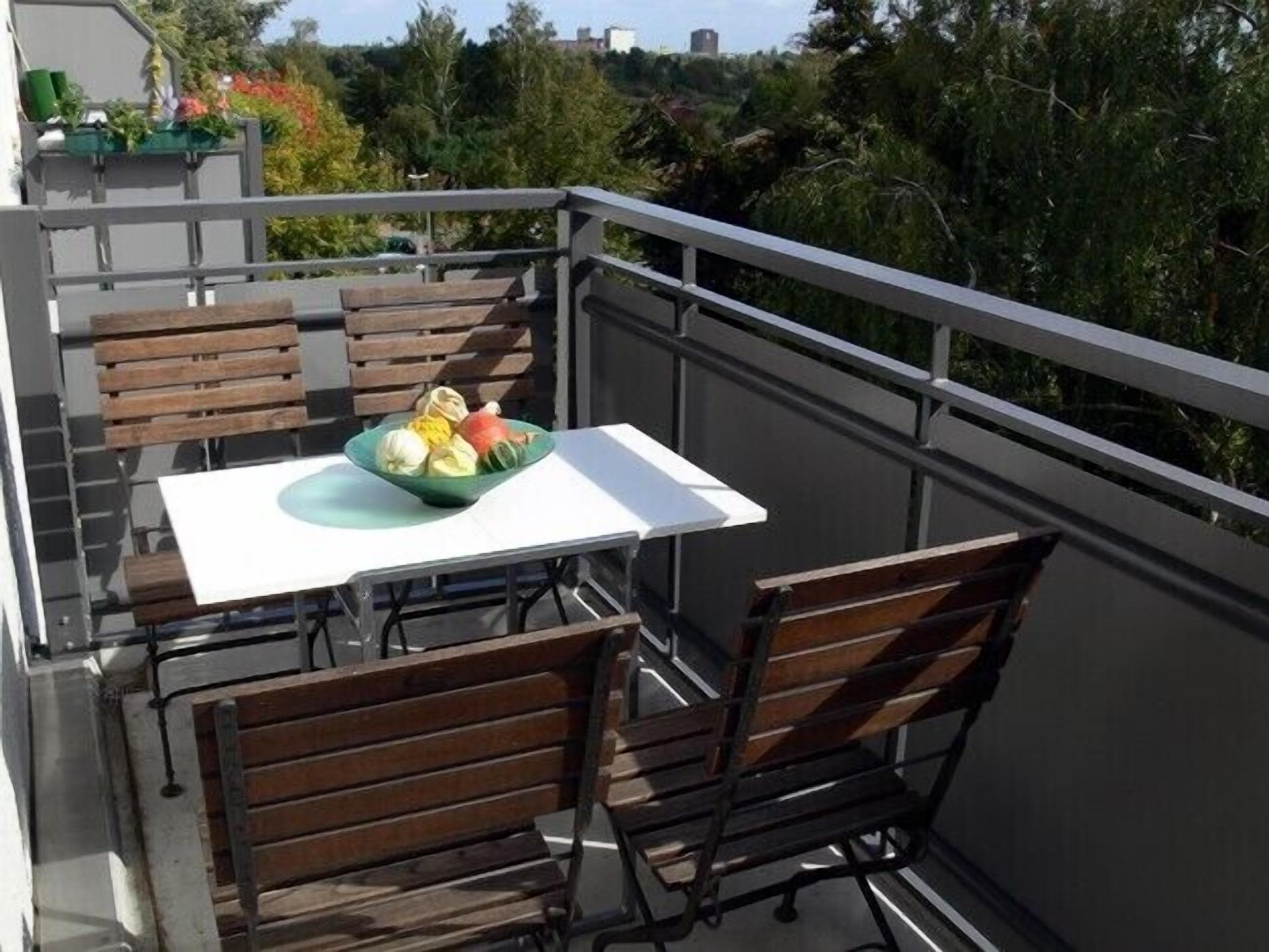 Ein Balkon mit Tisch und Stühlen, einer Obstschale und Blick auf Bäume und Gebäude.