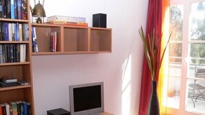 Ein Wohnzimmer mit Fernseher auf Holzfuß, einer Bücherwand voller Bücher, einem roten Schrank und einem großen Fenster mit Blick ins Freie.