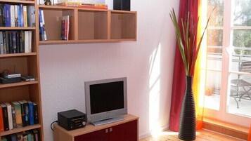 Ein Wohnzimmer mit Fernseher auf Holzfuß, einer Bücherwand voller Bücher, einem roten Schrank und einem großen Fenster mit Blick ins Freie.