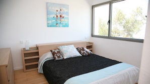 A bedroom with a bed, a nightstand, a wooden dresser, a painting on the wall, and a window with a view of trees. - MODERN villa with pool and fantastic sea views.  (PUERTO DE SOLLER, MALLORCA)
