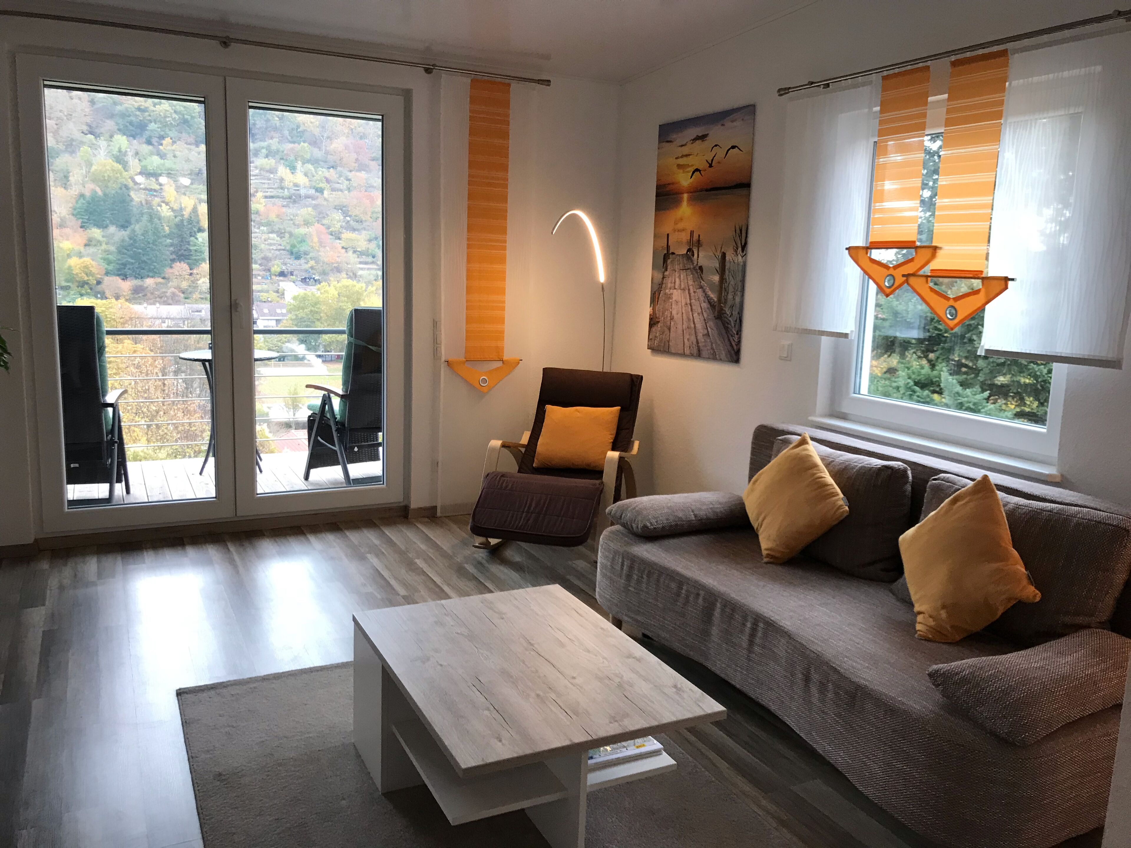 Un salon moderne avec un canapé, une table basse et un fauteuil. Une grande fenêtre, ornée de rideaux, offre une vue sur les arbres.