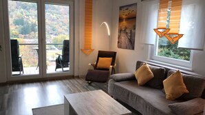 Uma sala de estar moderna com um sofá, uma mesa de centro e uma poltrona. Há uma janela grande com cortinas e vista para árvores.