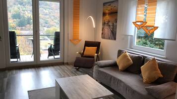 Ein modernes Wohnzimmer mit einer Couch, einem Couchtisch und einem Sessel. Ein großes Fenster mit Vorhängen bietet Blick auf Bäume.