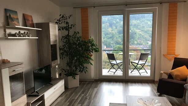 Uma sala de estar moderna com televisão, uma planta e uma varanda com cadeiras.
