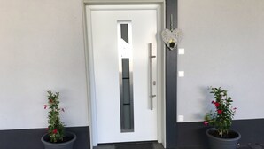 Uma porta branca com puxador vertical, ladeada por dois vasos de plantas com flores vermelhas, e uma decoração em formato de coração na parede.