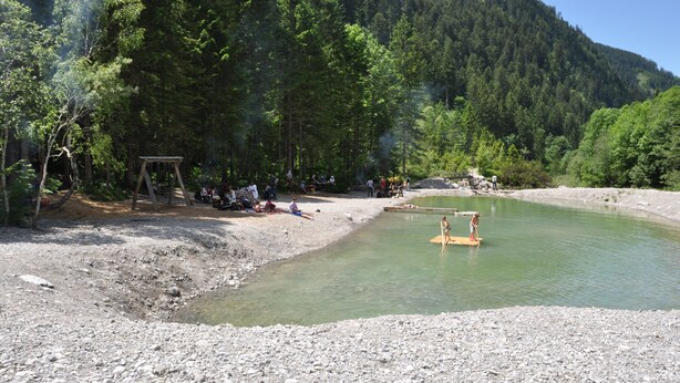 Un entorno natural con un río, un columpio de madera y gente relajándose junto al agua.