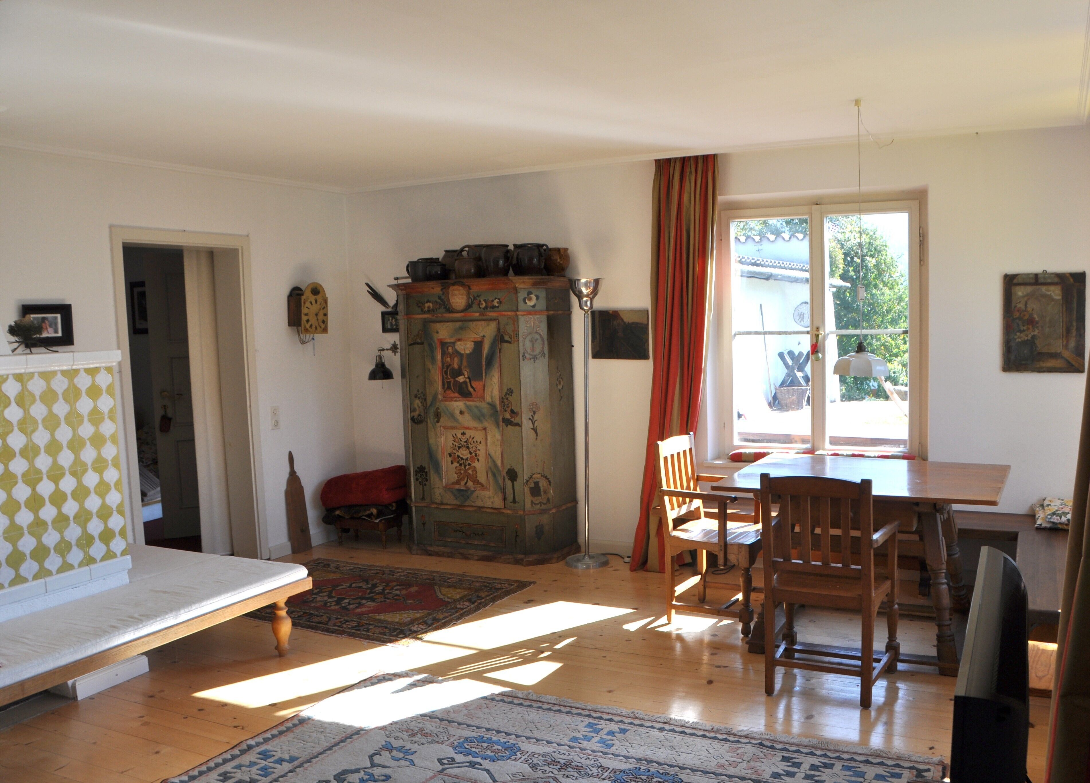 A room with a wooden table, chairs, a cabinet with artwork, a bed, and a window with curtains.