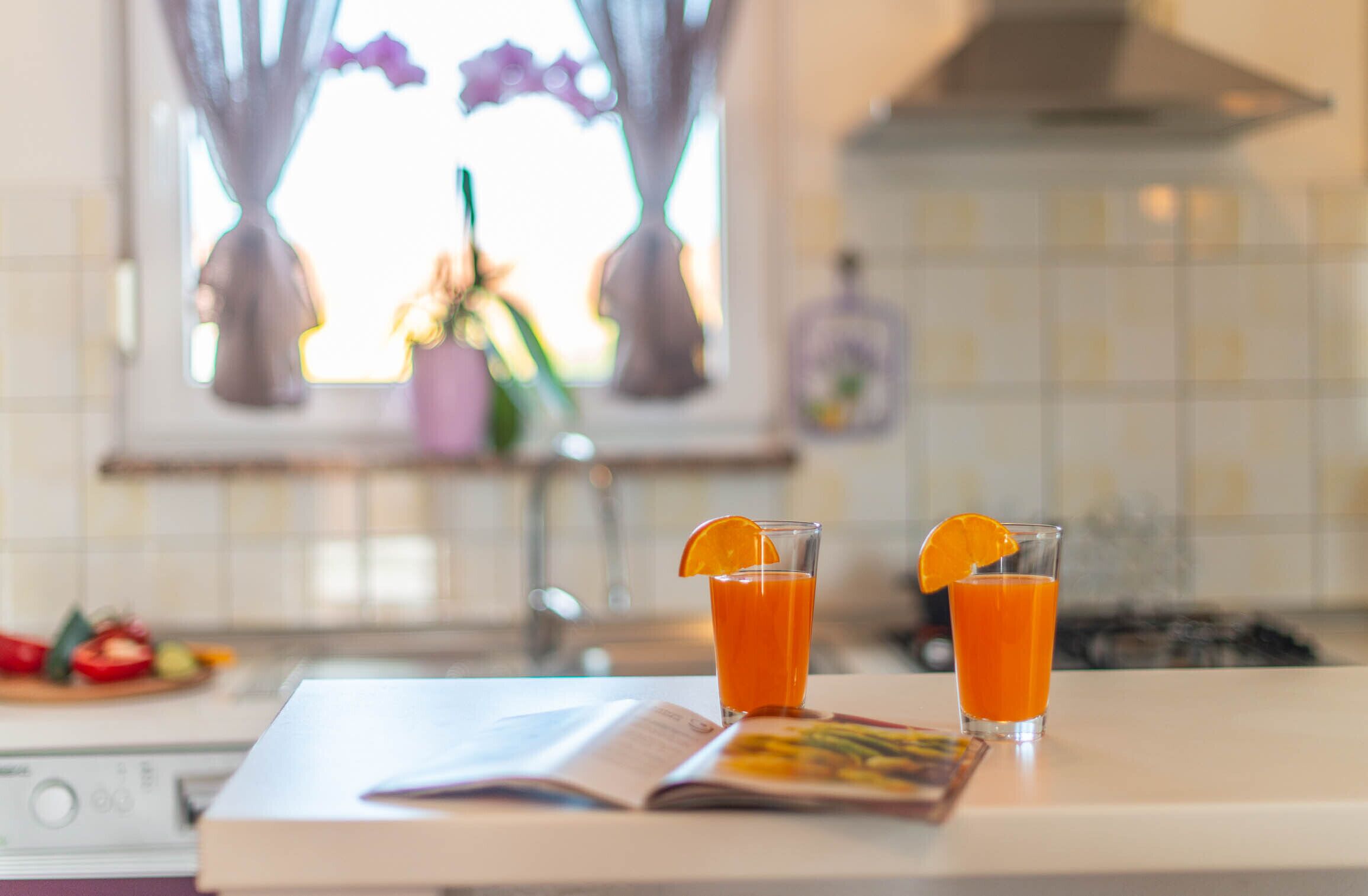 A kitchen with two glasses of orange juice on the counter, a book, and a window with curtains.