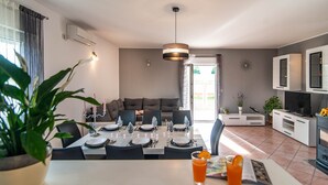 Een moderne eetruimte met een tafel voor vier personen, een bankje op de achtergrond en een keuken met een televisie en kasten.