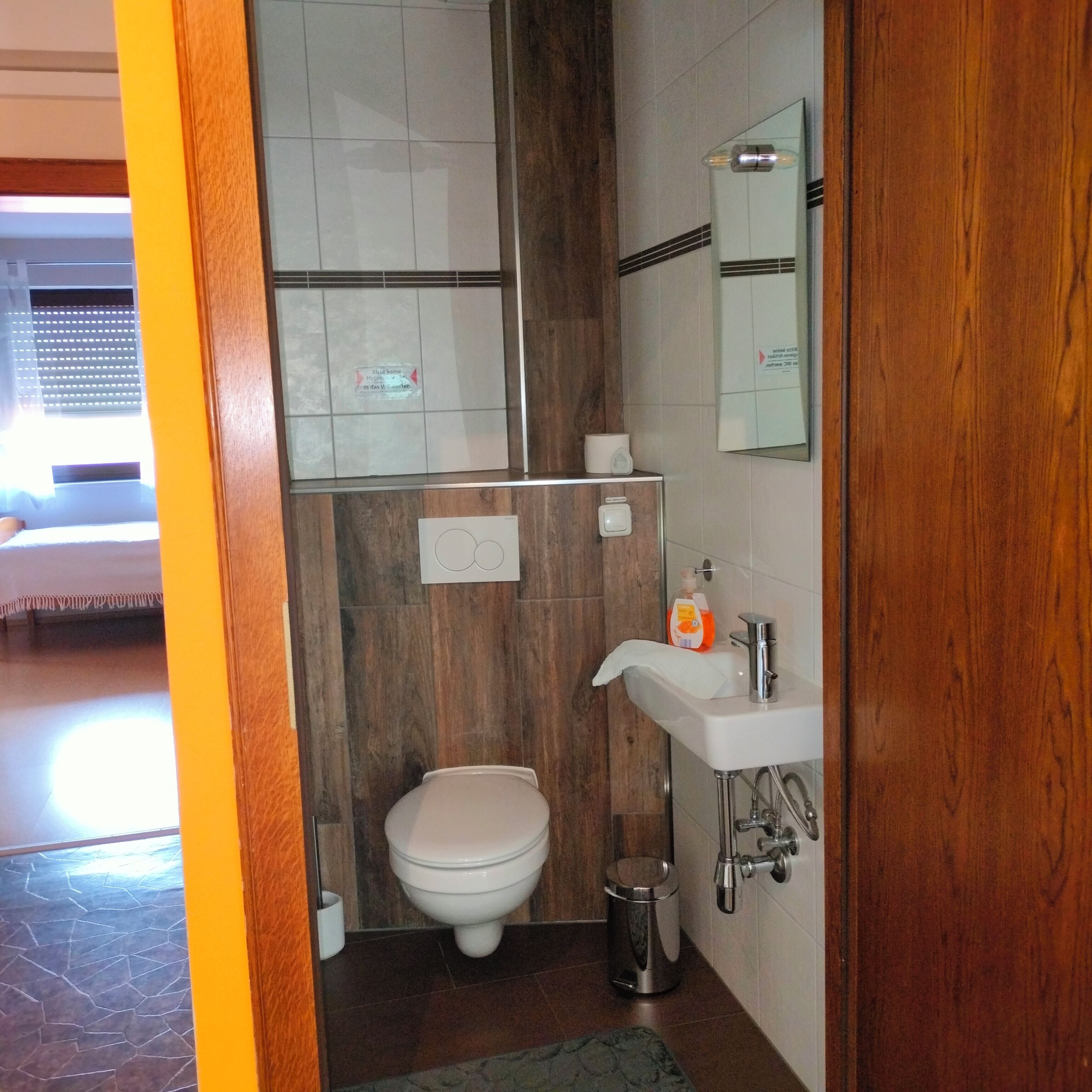 A bathroom with a toilet, sink, and mirror. There is a trash can and a bottle of soap. The walls are tiled, and there is a wooden panel on one side.