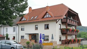 A two-story building with a red-tiled roof, a balcony with flower boxes, and a sign indicating it is 1 km from Hundsbach. - Luxurious, spacious **** apartment on a farm - family friendly (Tann (Rhön))