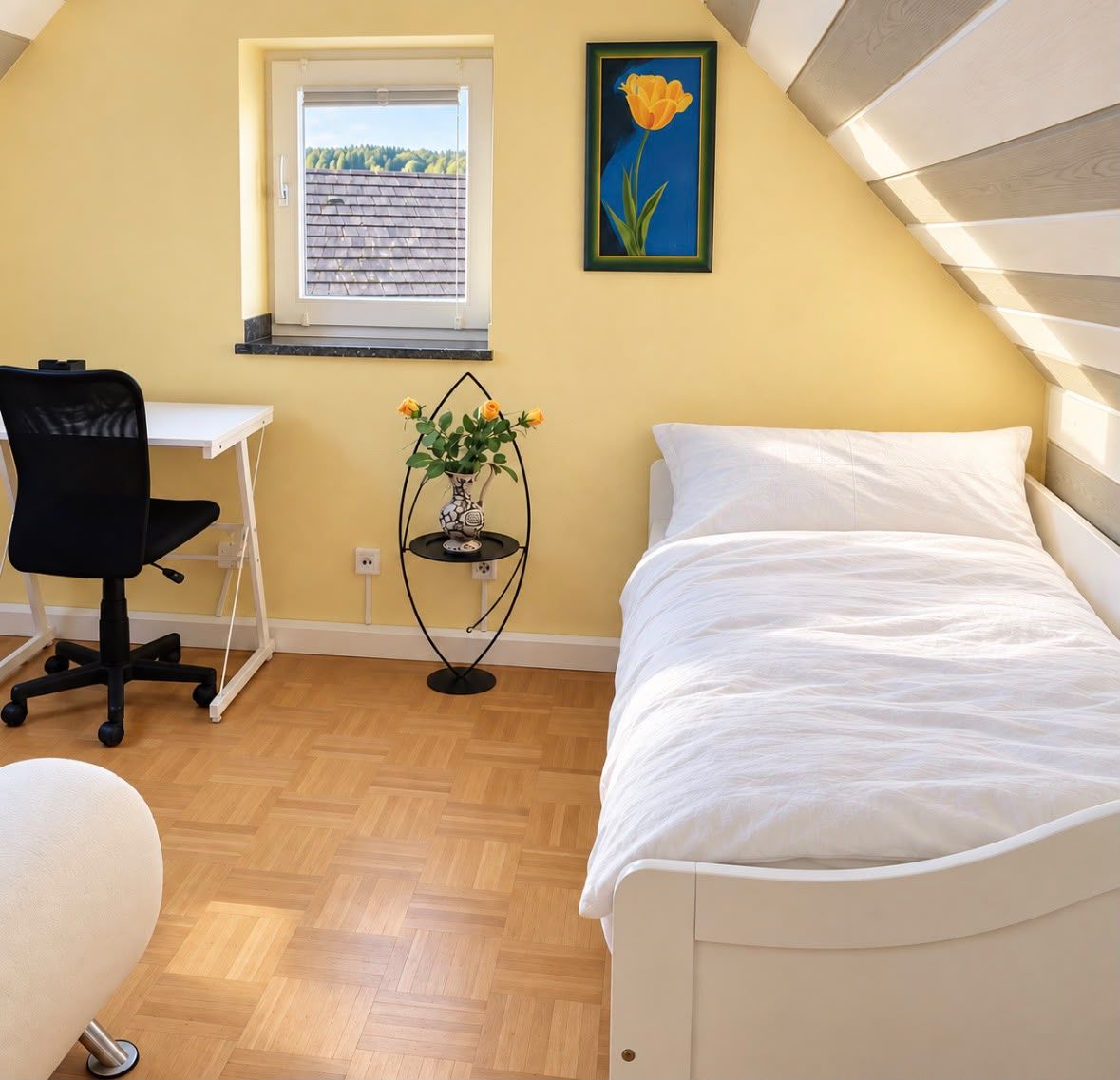 A bedroom with a bed, a desk, a chair, a small table with a vase of flowers, and a window with a view of trees.
