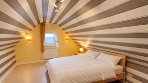 A bedroom with a sloped ceiling, a bed with white bedding, a nightstand with a lamp, and a window with curtains.