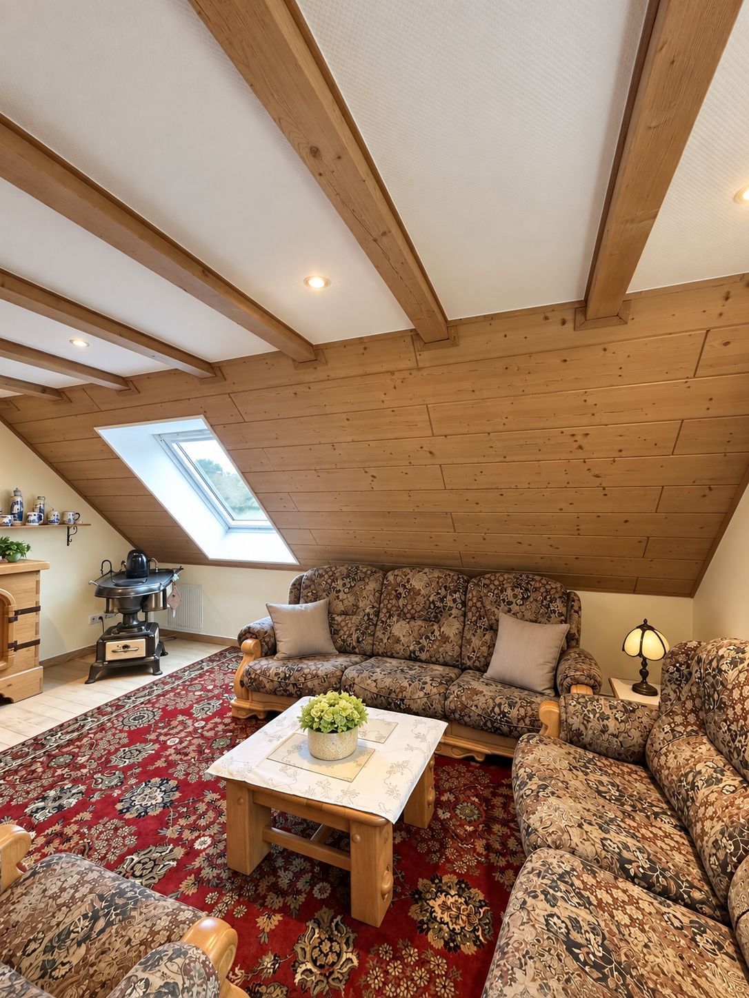 A cozy loft-style room with a sloped ceiling, wooden beams, a large sofa, a coffee table, and a radiator.