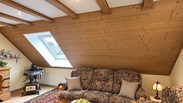 A cozy loft-style room with a sloped ceiling, wooden beams, a large sofa, a coffee table, and a radiator.