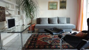 Una sala moderna con una mesa de centro de vidrio, un sofá gris con cojines, un sillón de cuero negro, una planta grande en maceta y dos cuadros en la pared.