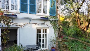 Un edificio blanco con contraventanas azules, un pequeño patio con mesa y sillas, y un jardín con plantas en macetas y un muro de piedra.