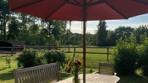 A wooden table and chairs under a large umbrella in a grassy outdoor area with a fence and trees in the background. - Vacation accommodation "Verwalterhaus" with view of the horse pasture -Gut Wittmoldt- (Wittmoldt)